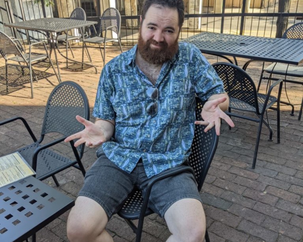 a white man with brown hair and a red beard sits sideways in a black metal chair with one leg under the armrest. He is wearing a printed blue button up short sleeve shirt with gray shorts. He is shrugging and he looks uncomfortable.