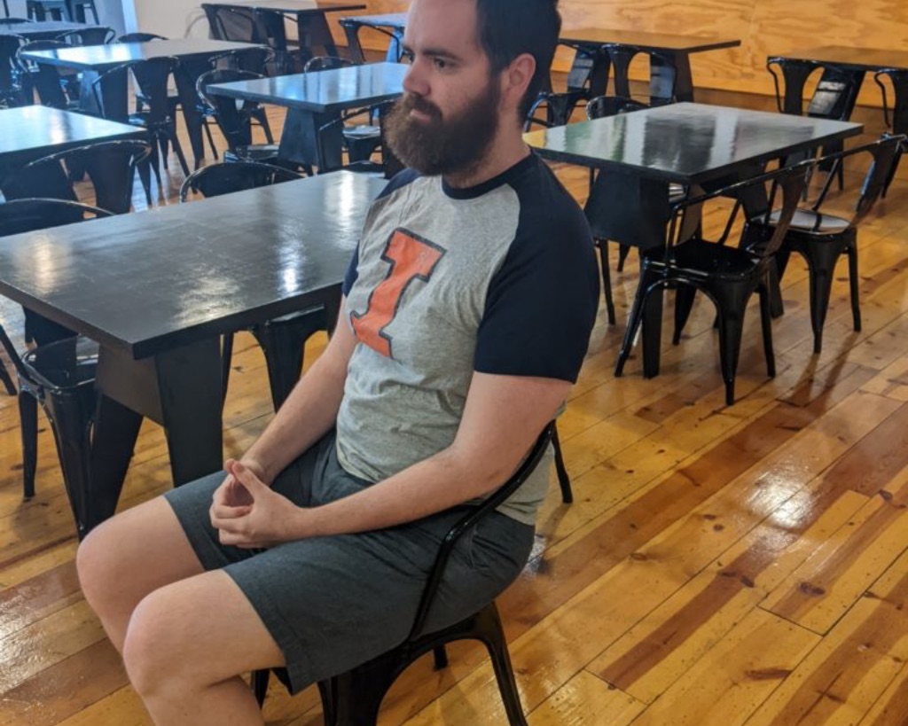 a white man with brown hair and a brown beard sits snugly in a black metal chair. He is wearing a gray t-shirt with navy sleeves and an orange I and gray shorts. He looks uncomfortable.