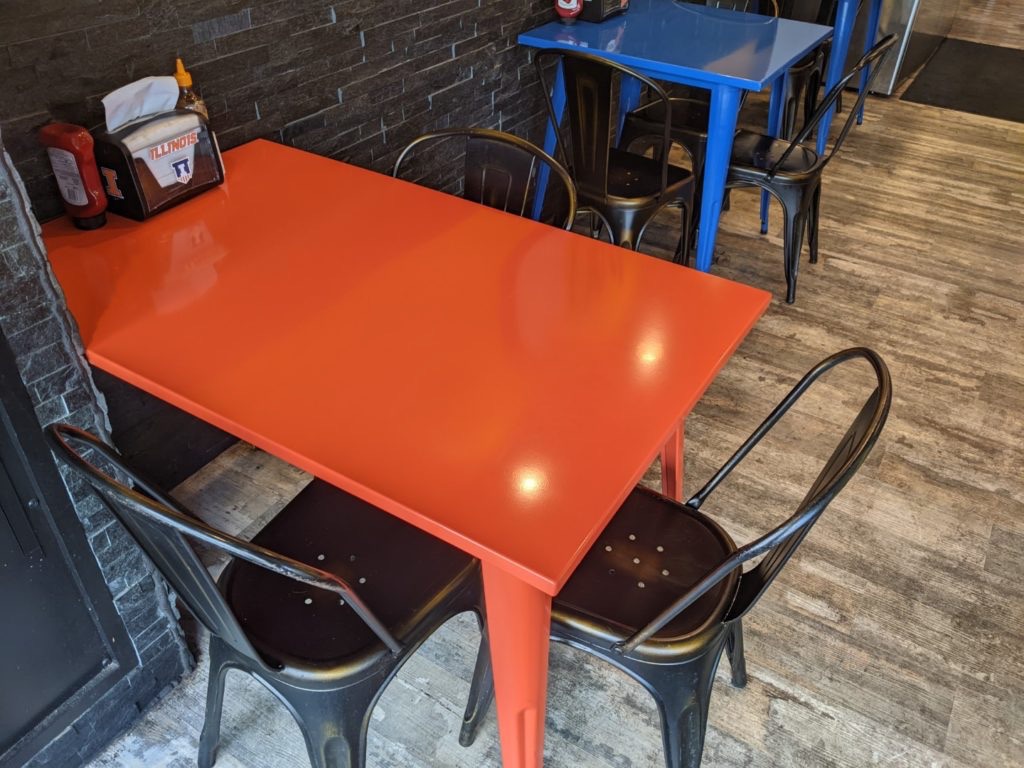 A picture from above of black metal chairs with curved backs sitting under a bright orange table. The floor is wooden and there is a blue stool in the background. 