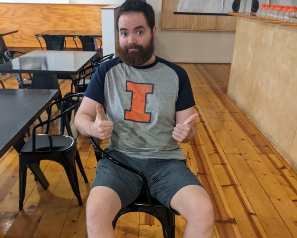 a white man with brown hair and a brown beard sits sideways in a black metal chair with one leg under the armrest. He is wearing a gray t-shirt with navy sleeves and an orange I and gray shorts. He is smiling and giving a thumbs up.