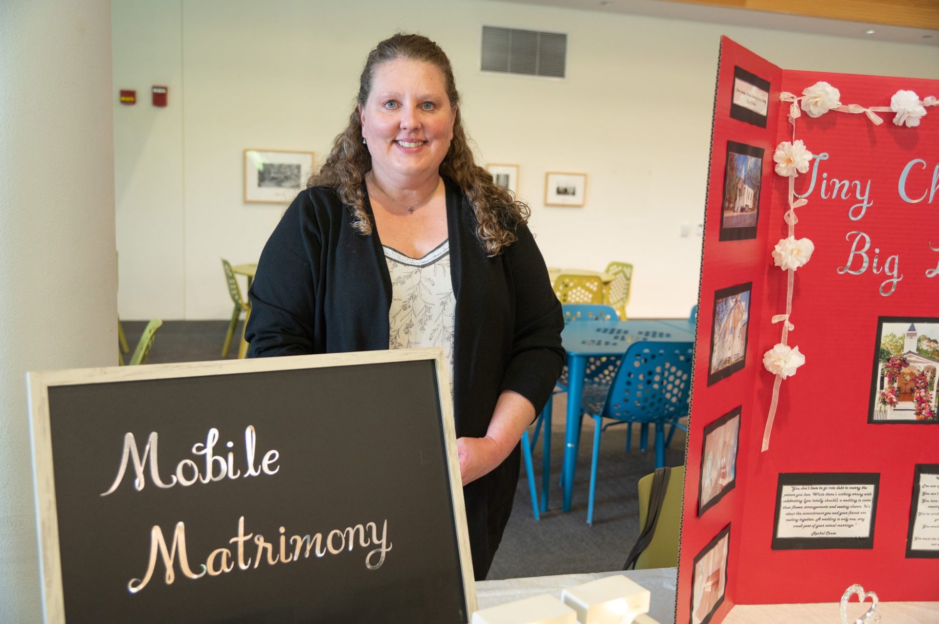 A white woman with long light brown hair, wearing a white shirt and black sweater, is standing behind a white table. There is a red tri-fold board on one side, of the table, and a black sign that says Mobile Matrimony in script on the other side.