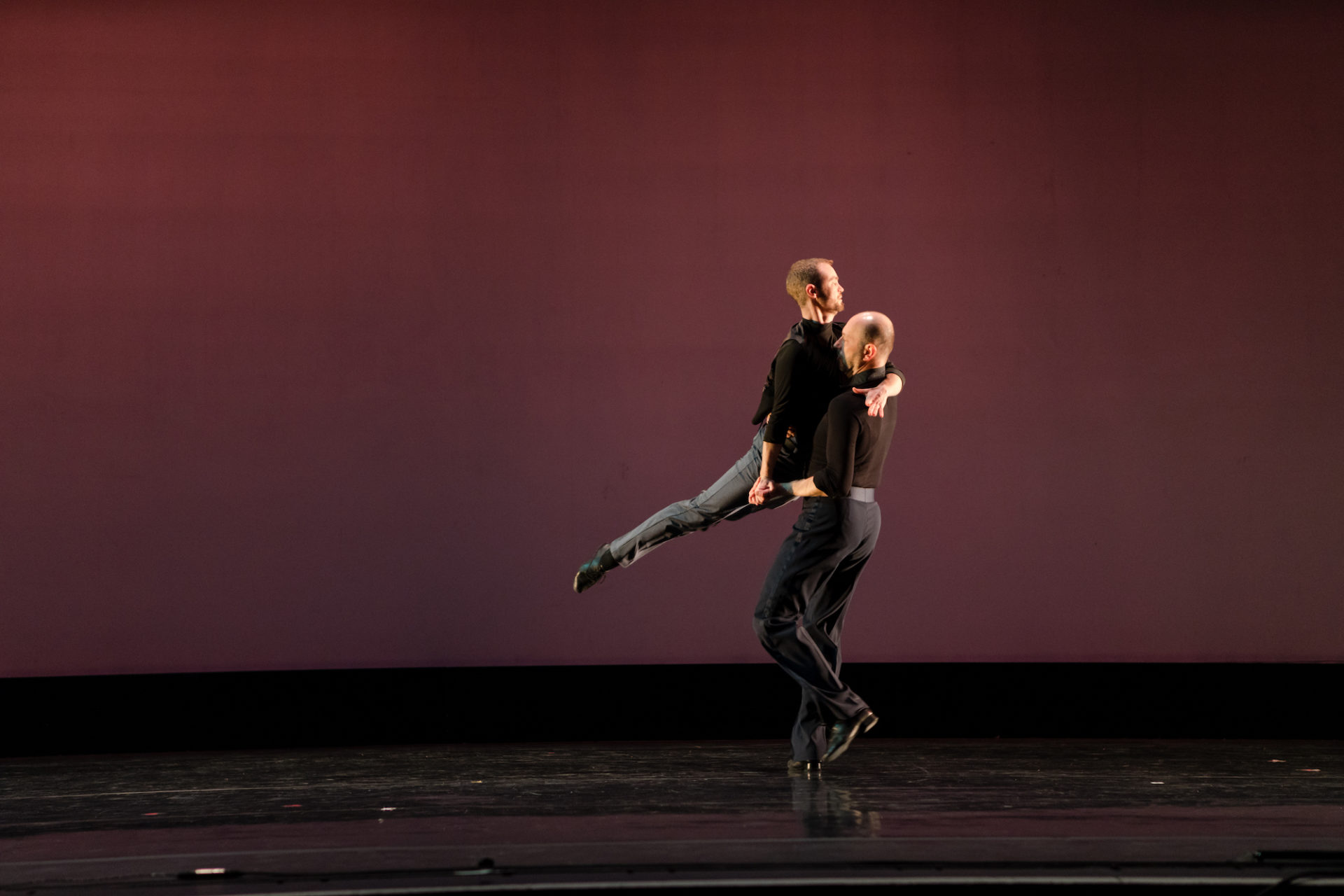 Alex Tecza and Kato Lindholm dance on stage at The Virginia. They wear matching grey pants and black shirts. Tecza is holding Lindholm as they spin around with Lindholm's leg extending out.