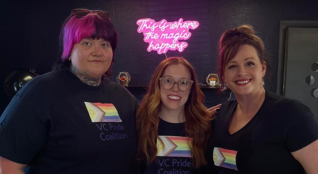 Three women stand together in front of a neon sign that says "this is where the magic happens" smiling at the camera. They are wearing black t-shirts with a pride flag on them.