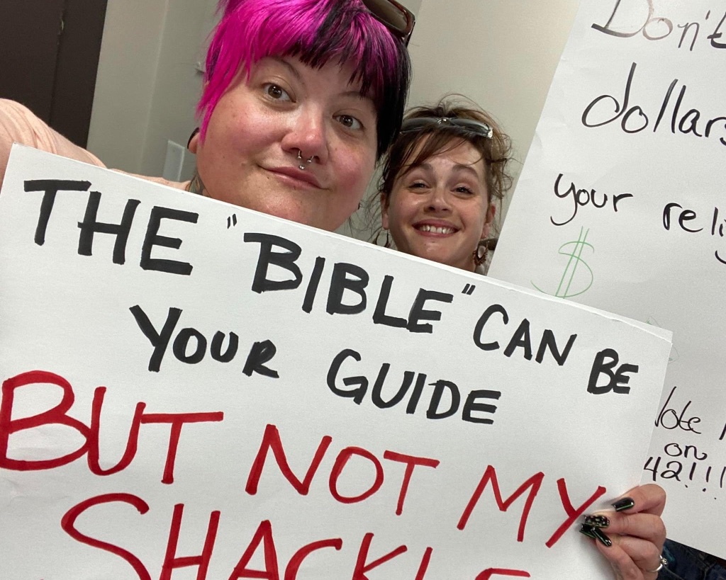 Two white women are holding signs, one reads "the Bible can be your guide but not my shackle" 