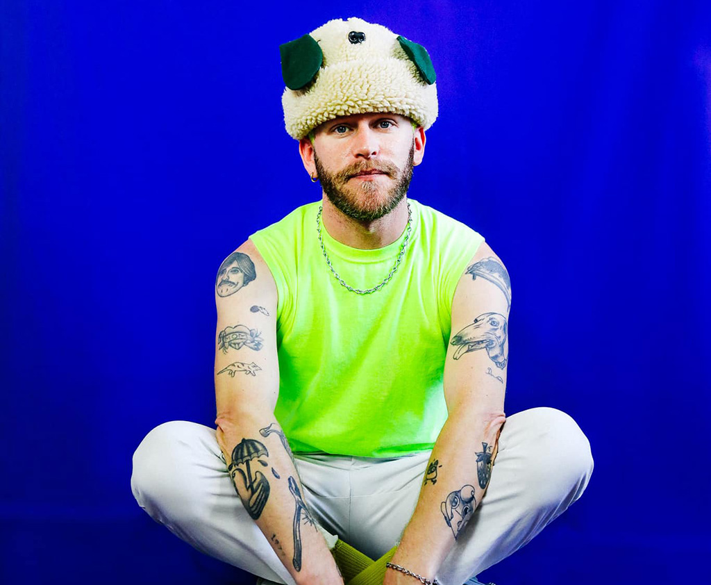 Music artist Gay Meat clad in a puppy hat, sleeveless bright neon t-shirt, with his legs crossed