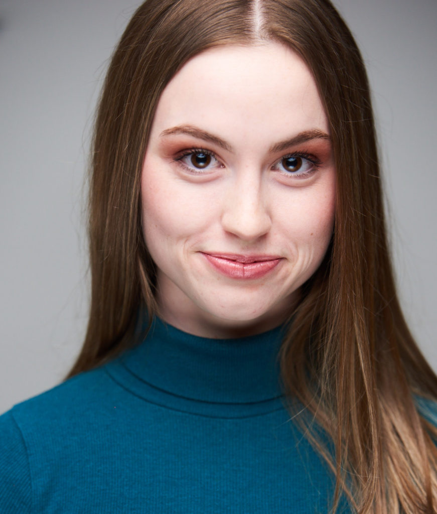 A headshot of Haley Gillespie. A young white woman with long brown hair parted down the middle. She looks straight at the camera with a smile. She is wearing a blue-green turtleneck. 
