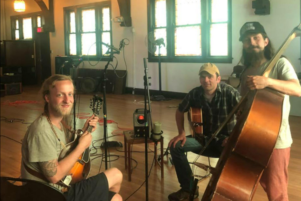 Three members of Hobnob String Band in a wide open recording space with recording equipment around, holding their instruments and looking into the camera.