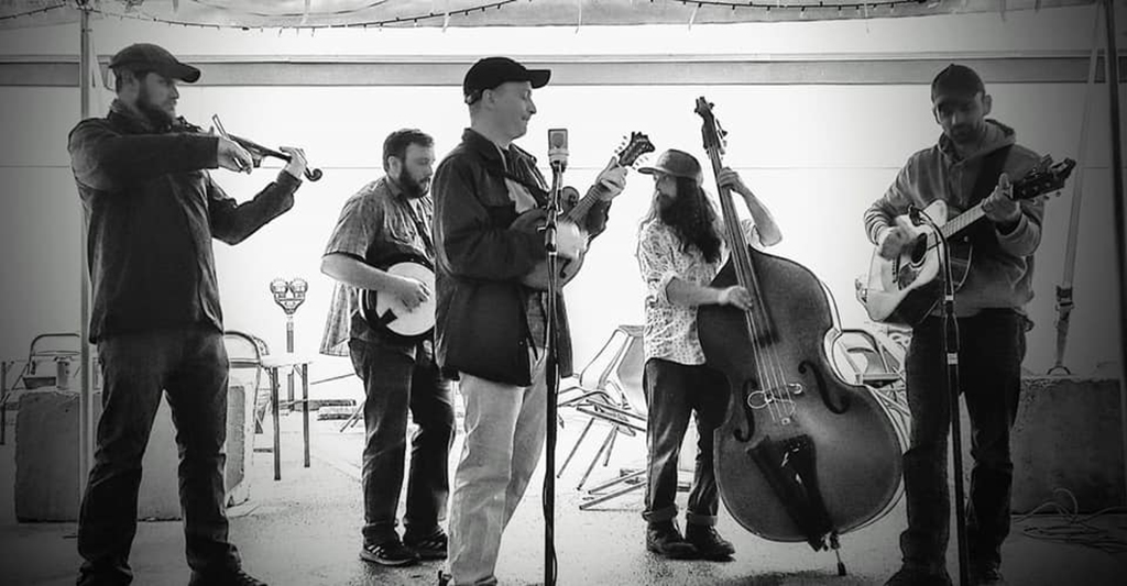 This image shows a group of seven people playing musical instruments in an indoor setting. There are four men and three women, all wearing casual clothing. The instruments being played include a guitar, cello, bass fiddle, and banjo. All the musicians appear to be performing together as part of a musical ensemble or band. The dominant colors in the image are grey, black and white.
