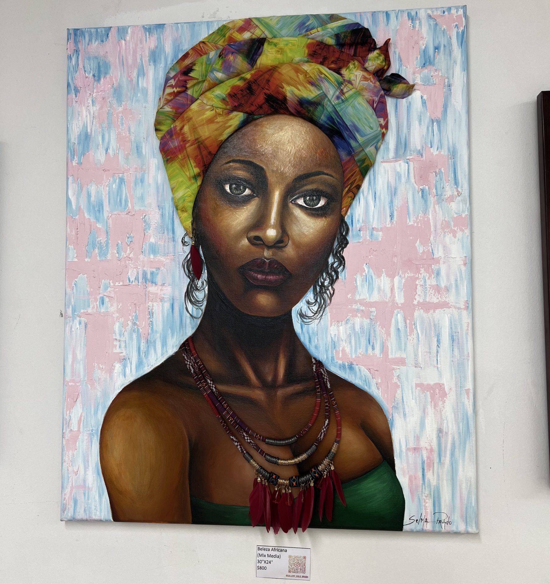 Mixed media painting by Selma Prado. A front view bust of a black woman wearing a green strapless top and a colorful turban. The turban and necklace are actual materials placed on the canvas. The background is abstract brush strokes of light pink and blue.