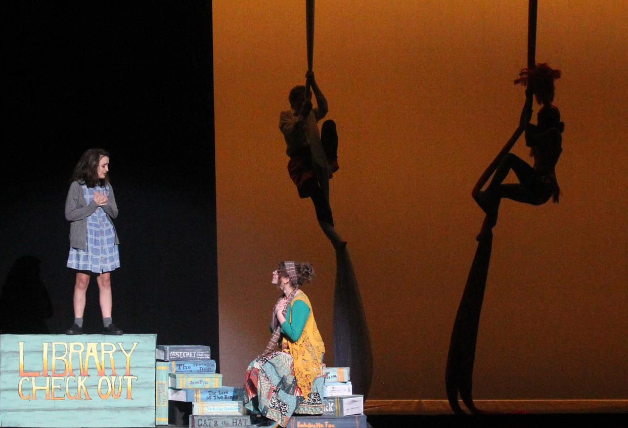 Two actresses appear on stage. One sits on a stack of books and looks up at another actress who is standing on a large sign that says "Library Check Out". There are two people suspended mid air on long pieces of fabric in the background.
