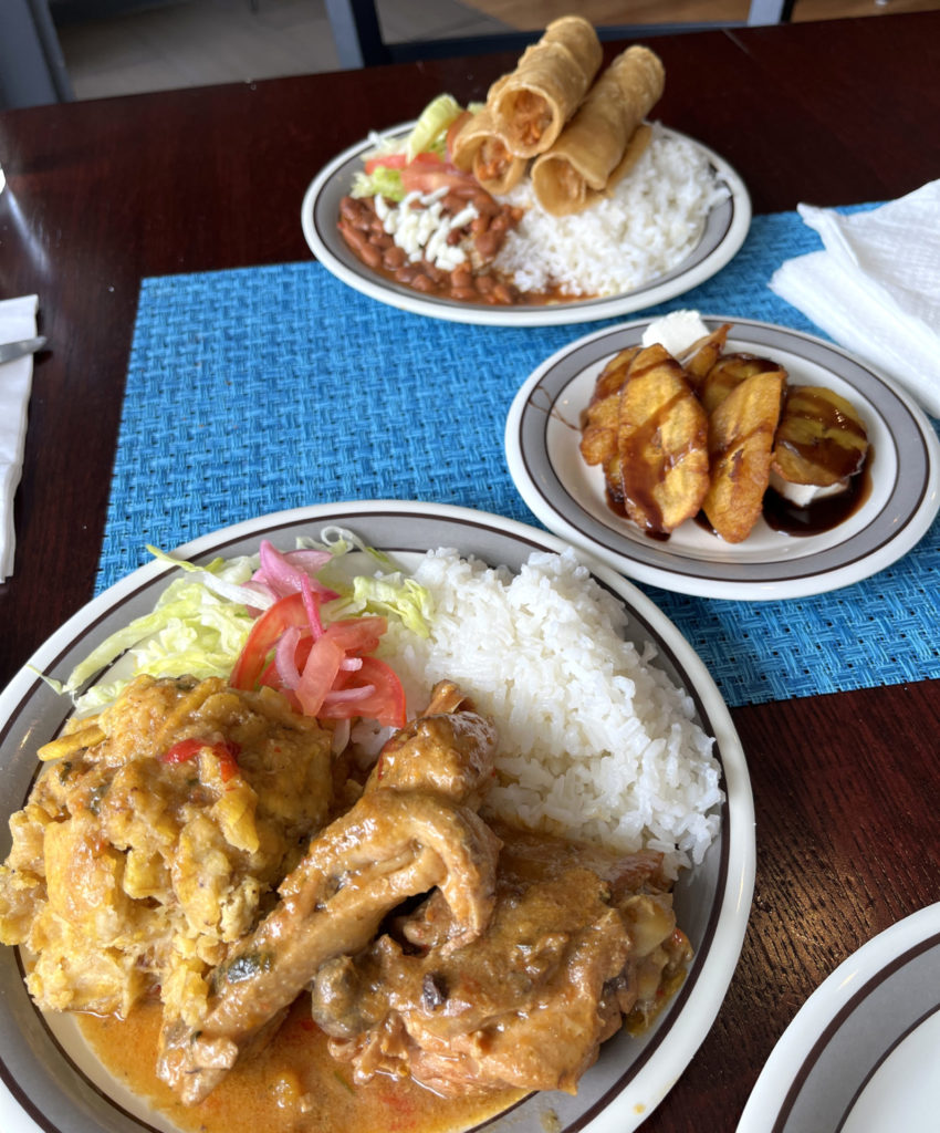 Three dishes on a dark wood table with a blue placemat at El Paraiso. Chicken mofongo with white rice; fried sweet plantains; three flautas with white rice and beans.