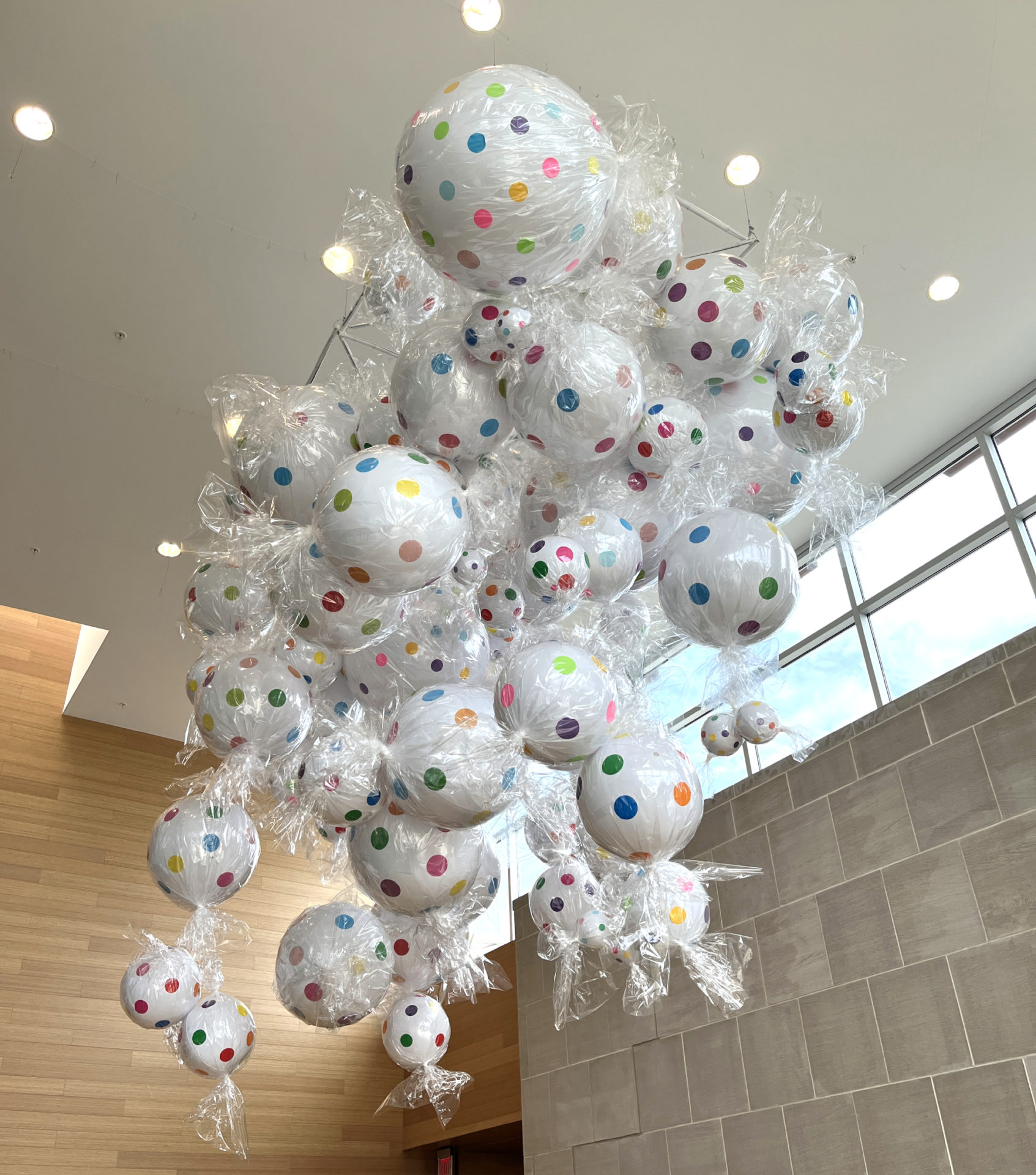 Installation hanging from the ceiling of the Champaign Public Library lobby. A lot of sculptures that are made to look like wrapped candies are large, white with colorful polka dots and clear wrappers.