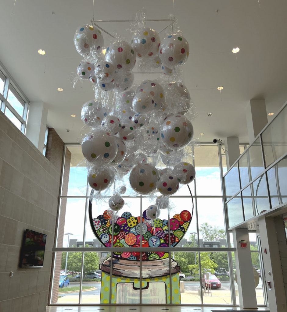 A view of the Champaign Public Library's lobby, viewed from inside looking out. An installation of large, fake candy in clear wrappers hangs from the ceiling. On the large glass window beyond is a painting of a gumball machine filled with colorful candies.