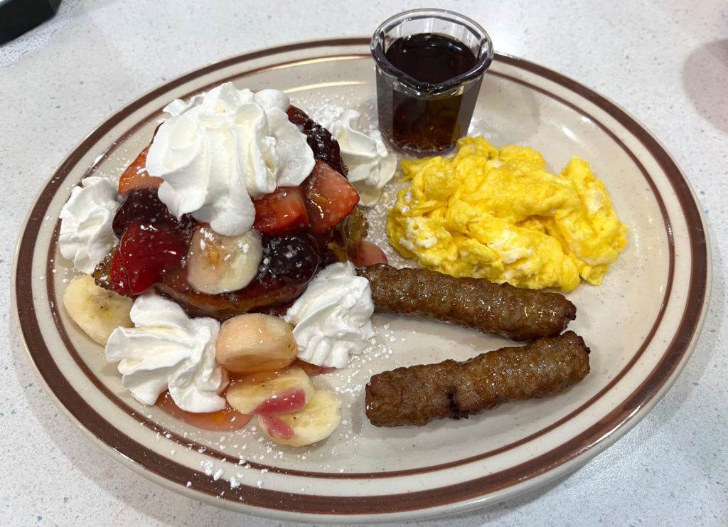 A plate of the homestyle cinnamon stuffed French toast fresh fruit combo at Sammy's Pancake House. An oval, white plate with brown trim is on a diner table. On the plate is a stack of French toast covered in fruit, glaze, and whipped cream, two sausage links, scrambled eggs, and a small container of syrup.