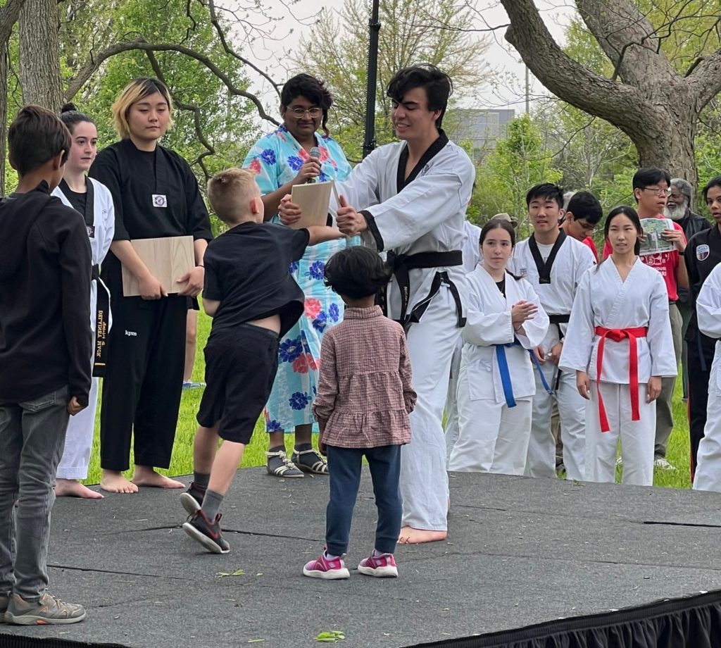 A young boy with blond hair is breaking through a wooden board, held by an adult in a white martial arts uniform and a black belt. There are several other people in uniforms standing near the stage.