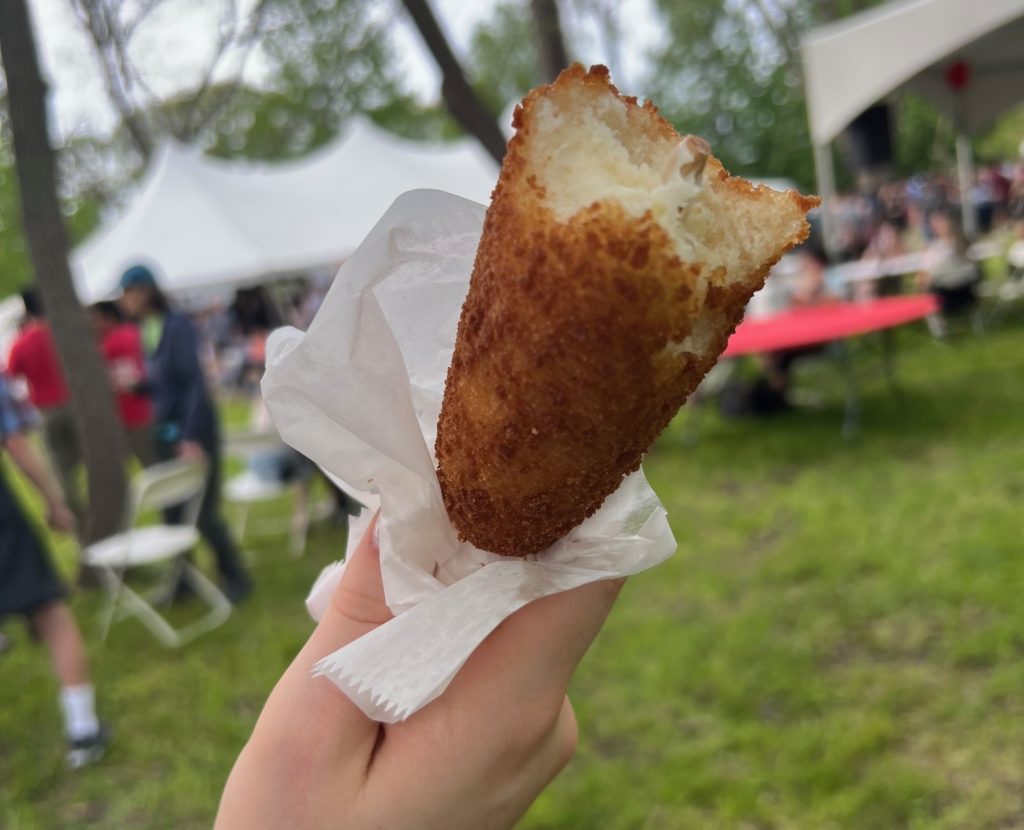 A friend corn dog with white tissue paper at the bottom. It has a bite taken out of it, showing gooey white cheese in the center.