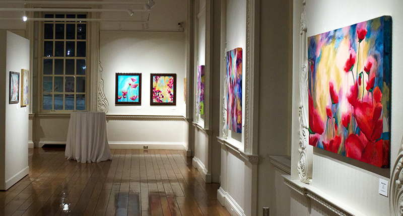 A portion of an art gallery with white walls, and several floral painting hanging. There are wood floors, and the lighting is casting a reflection of the paintings in the floor.