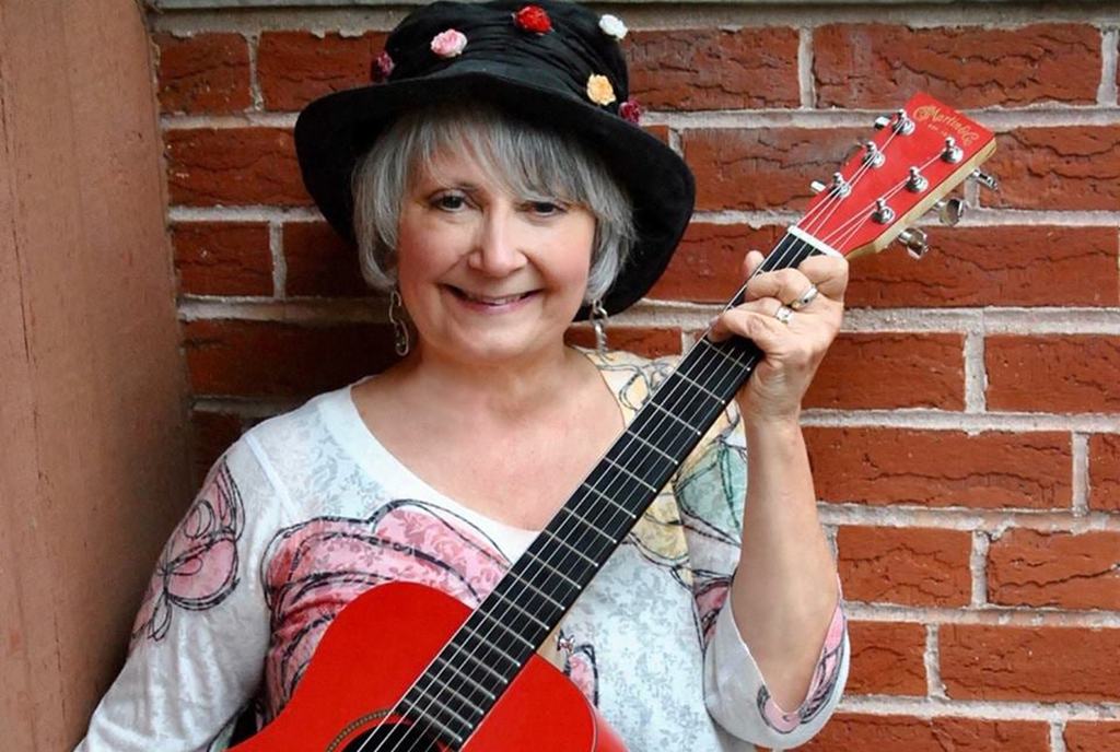 A woman with grey hair and a white large floral print blouse on with a black hat holding a guitar