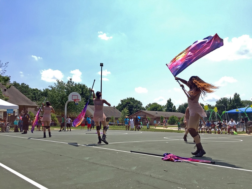 Two woman stand on a basketball court waving large flags in the air. There is a blue sky behind them and a few white clouds.