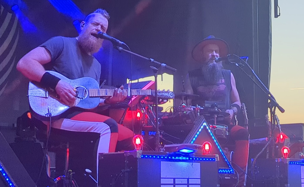 Two men with beards performing onstage, one with a guitar and the other playing electronic drums, both are singing into a mic