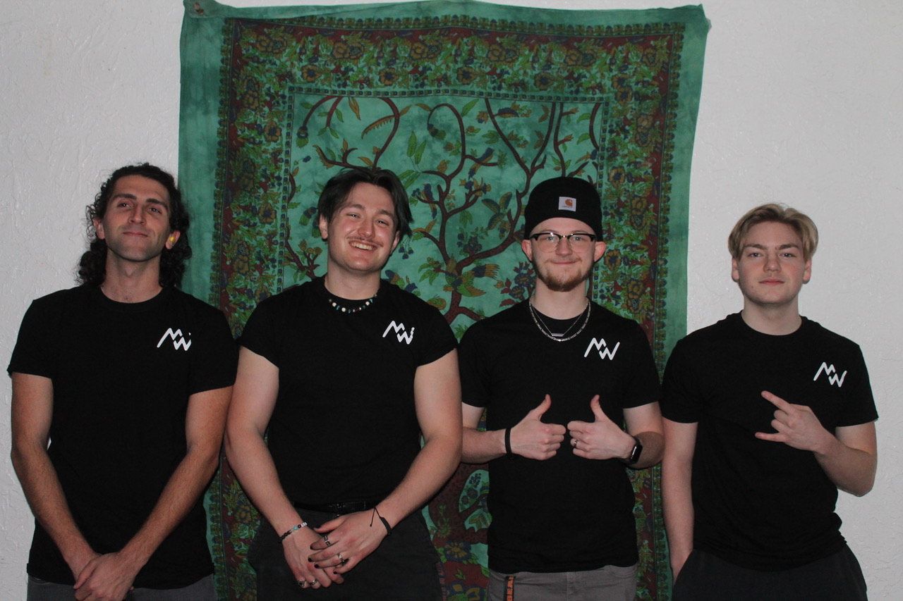 Four members of Manifest west, all wearing their own band's black t-shirts with their logo on it, standing in front of a green and brown tapestry pinned on an off-white wall.
