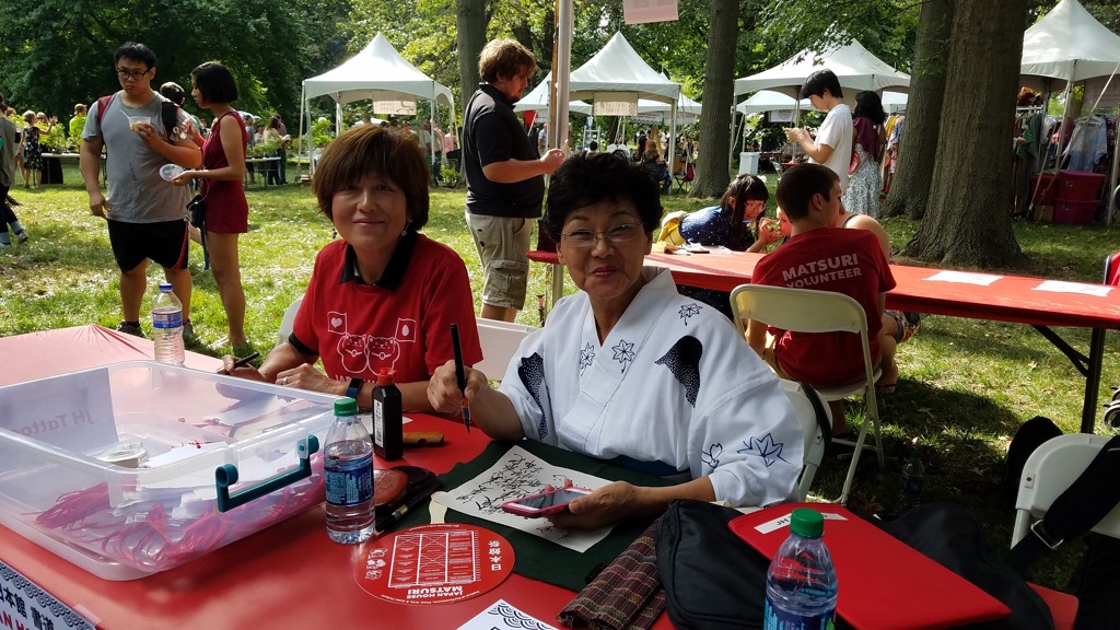Two women with short dark hair sit at a red table outside. There are people standing and white tents in the background. The woman on the right is wearing a white kimono with black star shapes, glasses, and is holding a phone and a paintbrush. The woman on the left wears a red shirt with a black collar. Both woman are smiling at the camera.