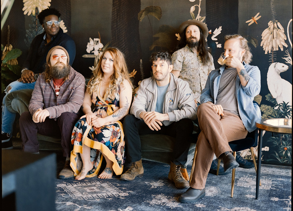 Six members of Merry Travelers either sitting on, sitting next to or standing behind a couch that looks like it's in a parlor. Three band members are looking into the camera. The band member sitting on the chair on the right is drinking something out of a cup.