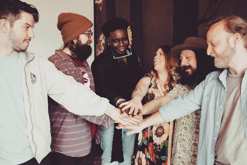 Five members of Merry Travelers standing in a half circle, all with one hand extended out in front of them. All of their hands are touching in the space in front. They are smiling, and most of them are looking at their drummer.