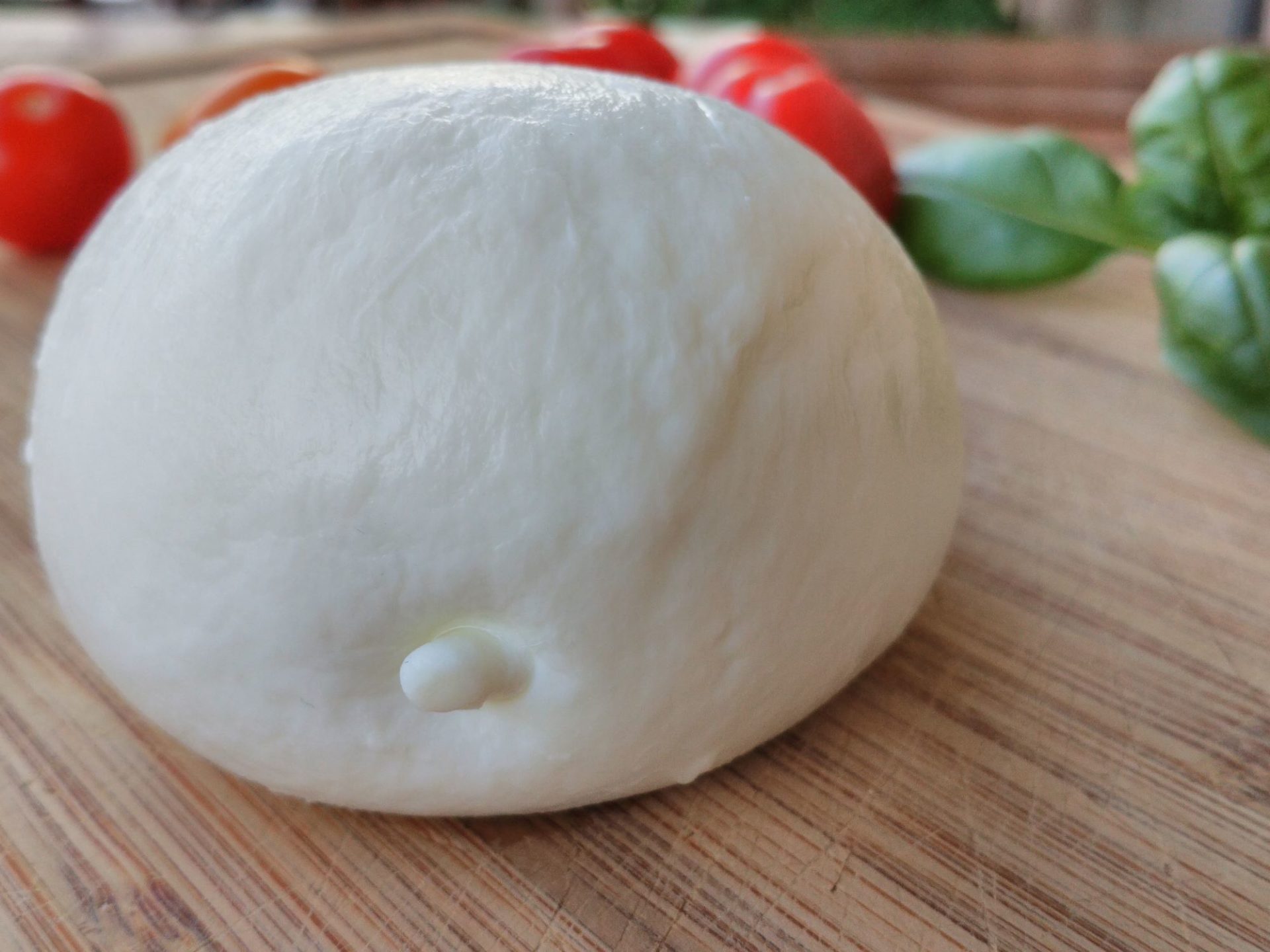 A ball of mozzarella cheese is siiting on a wooden counterop, with red tomatoes and green basil leaves in the background.