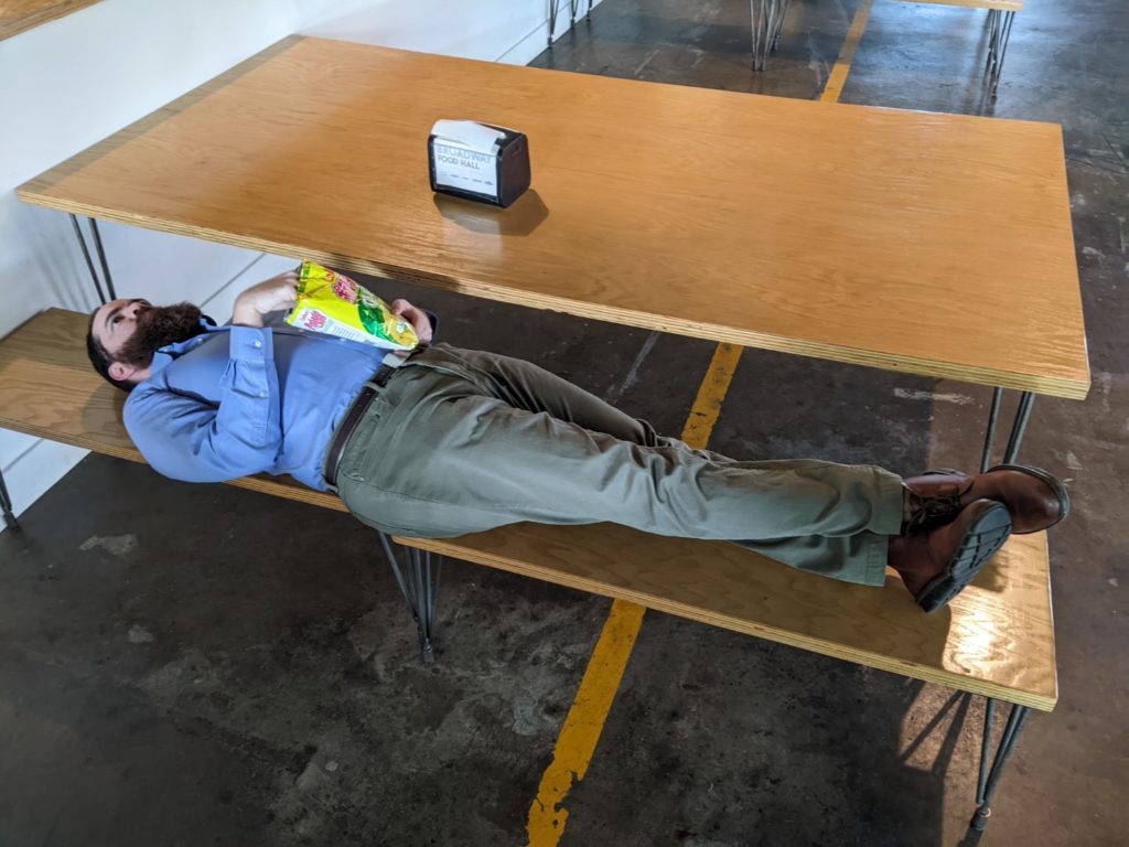 a white man with brown hair and a red beard lays on a wooden bench connected to a wooden table. He is wearing a blue button up long sleeve shirt and khaki pants. He has his ankles crossed and is holding a bag of chips.