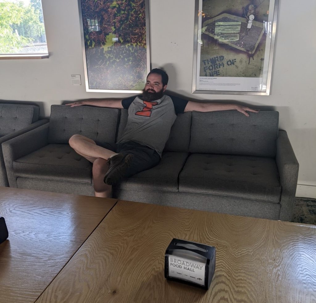 a white man with brown hair and a red beard sits comfortably on a grey couch. He is wearing a gray t-shirt with navy sleeves and an orange I and gray shorts. He has his right ankle up on his left knee and his arms on either side of the couch cushions.