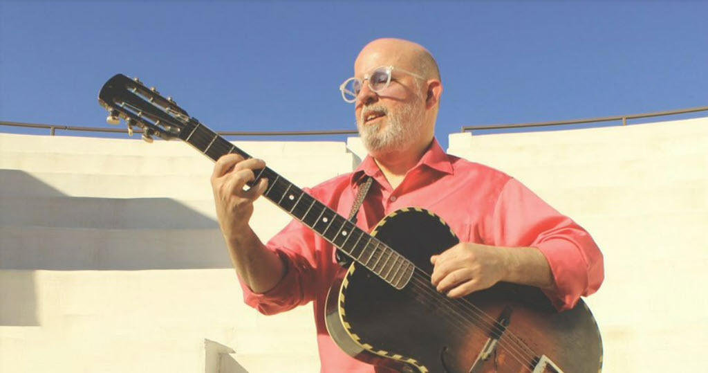 Man in a salmon short-sleeved shirt playing guitar out side with a high tan wall behind him.
