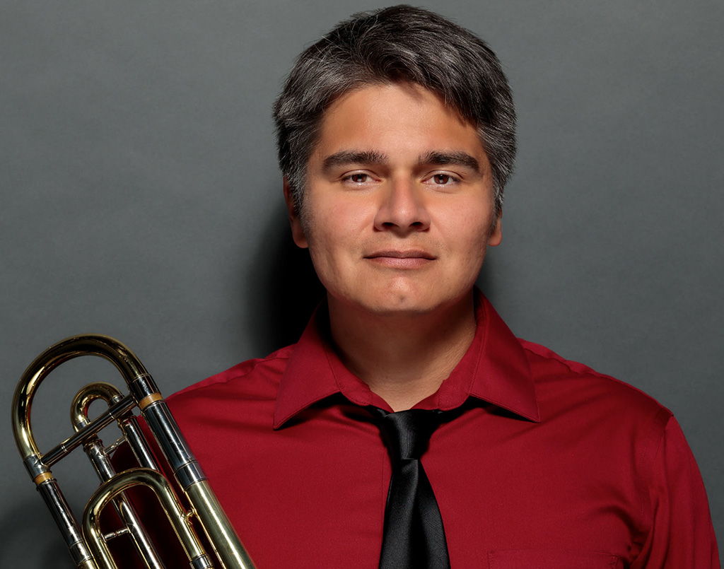 Close up on Peter Tijerina, wearing a red shirt, collar slightly unbuttoned with black tie on, holding a trombone.