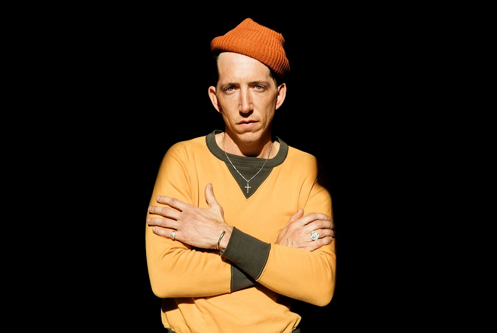 Pokey LaFarge in an orange knit cap, with a yellow shirt with olive green trim and a cross necklace.
