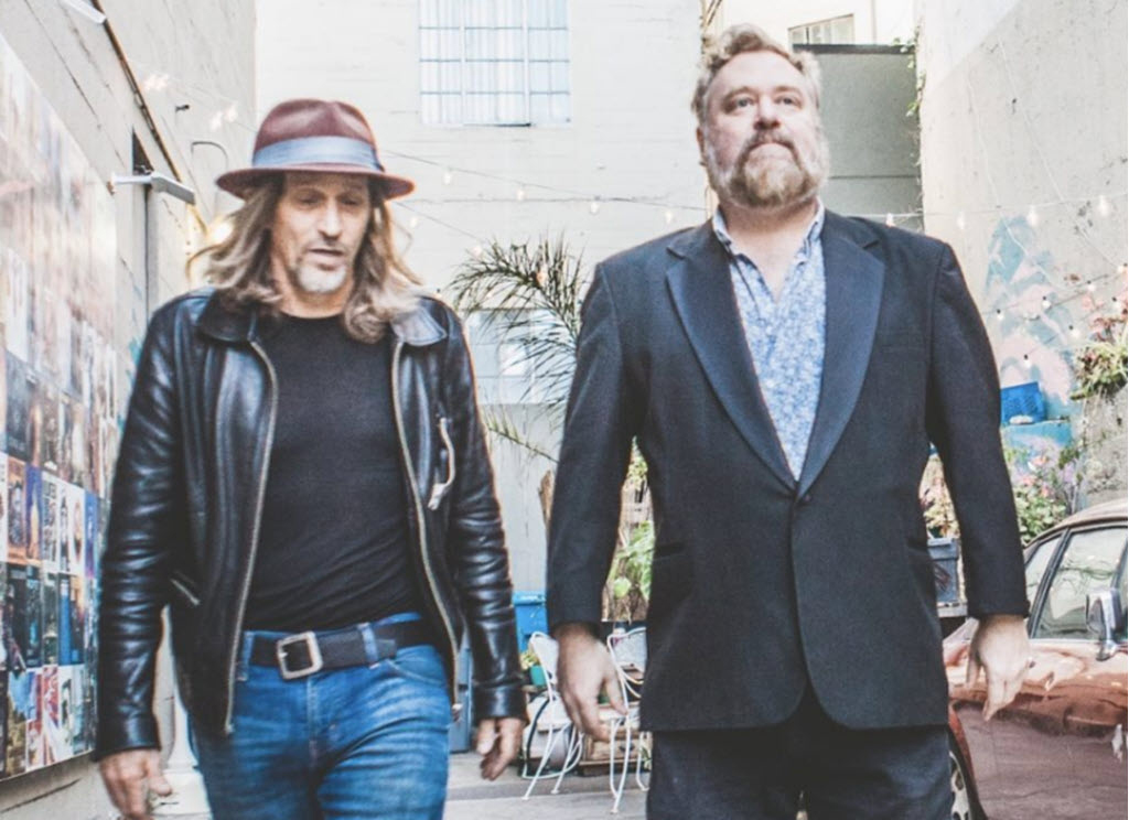 Two men next to each other. One appears to be walking and talking, and he's wearing a fedora, has long hair, a leather jacket, a black t-shirt, black belt and blue jeans. The man on the right is dressed in a suit jacket with a floral pattern shirt underneath. He is also wearing dress pants. He is tall with a reddish-grey beard and hair.