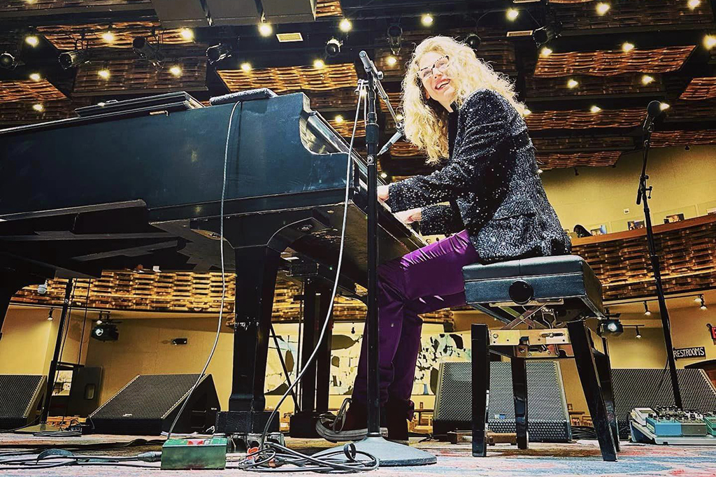 Photo of Sophie B. Hawkins with a black sequined jacket and purple pants playing a grand piano