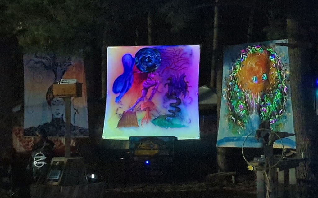 Three large psychedelic paintings sitting in front of a forest at night.