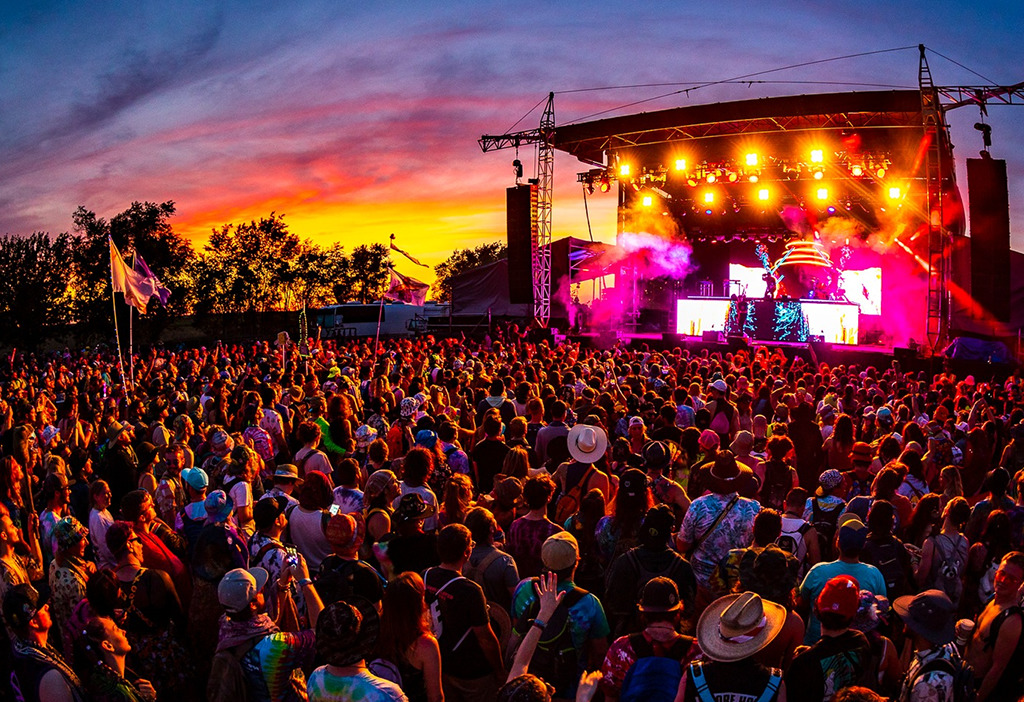 A photo of the crowd facing the stage with an EDM act performing. There are a lot of bright neon colors coming from the lights on the stage, and the sunset fills the sky in the background.