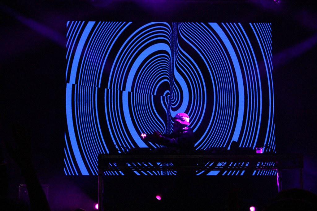 A EDM DJ behind a DJ table with a black and blue spiral graphic on a video screen behind him.