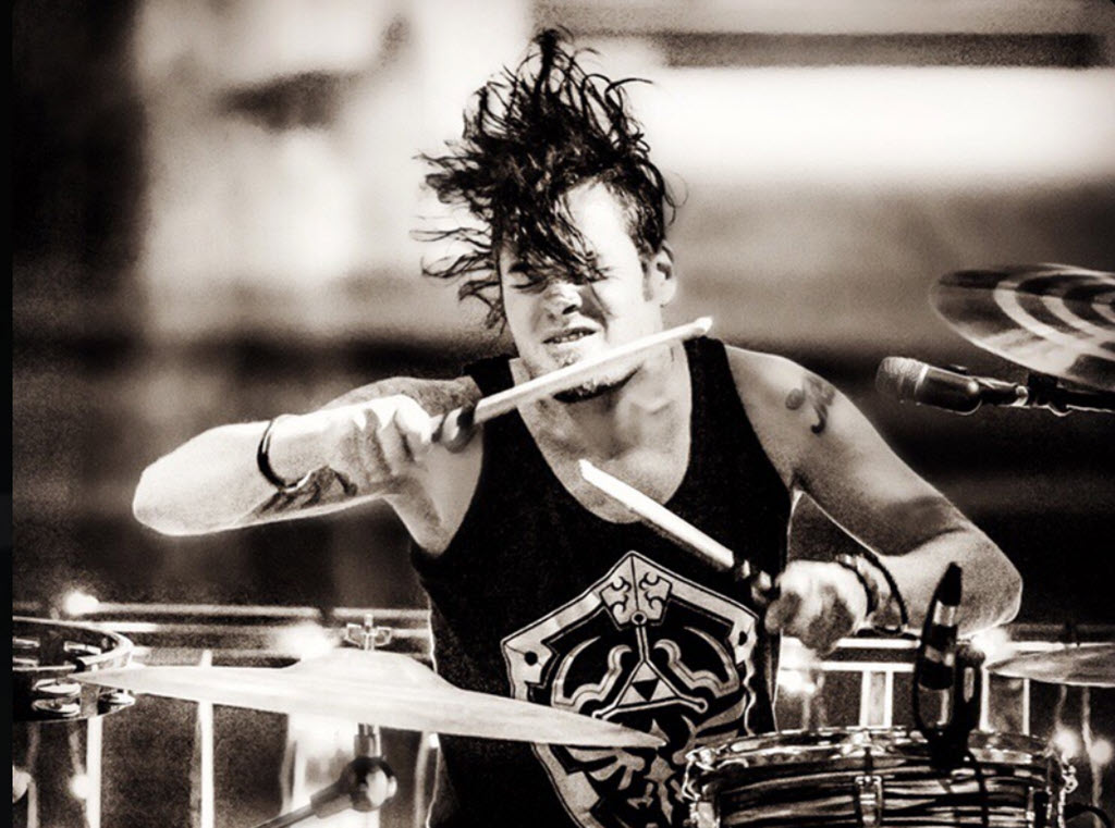 Black and white photo of a rock band drummer with his hair flying and his sticks poised to hit his drums. His eyes are closed and he is wearing a black tank top.
