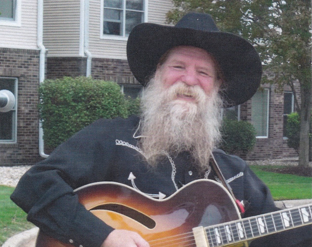 This image is of a man with a long beard and a black hat playing an acoustic guitar outdoors. He is wearing a cowboy-style hat, and his clothing appears to be mostly black in color.
