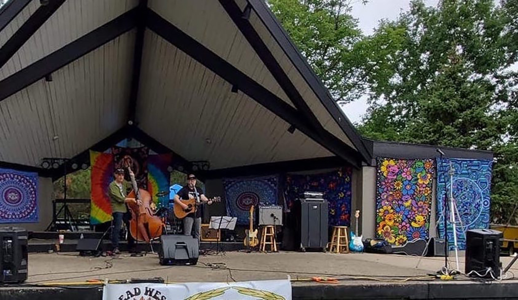 Far away shot of an upright bass player and a guitarist playing on an outdoor stage that has psychedelic tapestries hung in the background