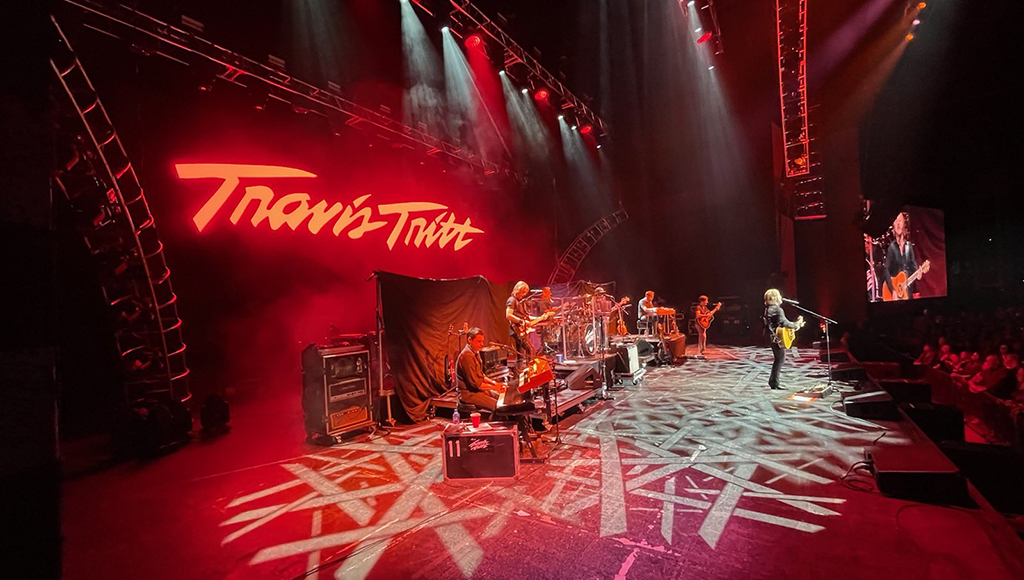A country band playing onstage. The stage is large and lit in red. The artist's name is on the wall behind the band.