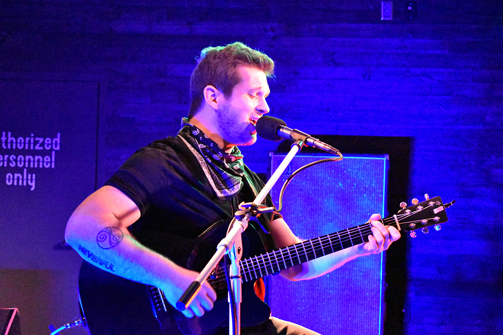 A man with a black t-shirt and bandanas around his next singing while playing guitar in blue light onstage.