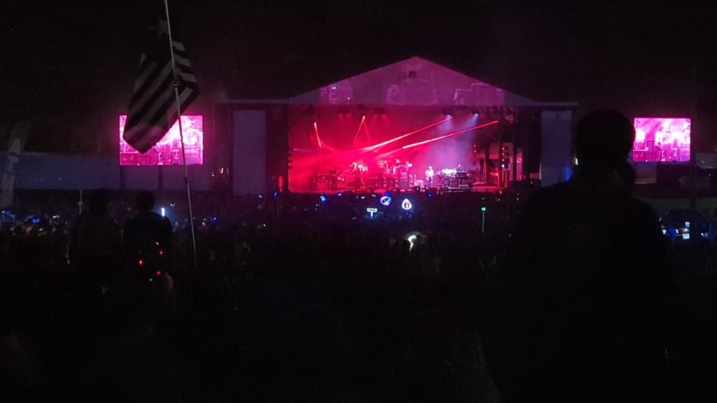 A far away view of a stage lit in red at night with a band on it. USA flag in the foreground and a large video screen on both sides of the stage.