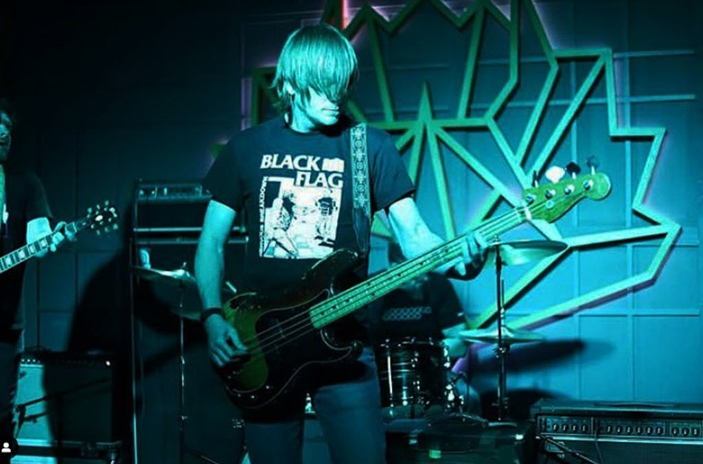This image is of a man playing an electric guitar on stage. He is wearing a black shirt and jeans, and has long dark hair. The background of the image is mostly dark with some blue lighting illuminating the stage. There are other instruments visible in the background, including a steel drum to his left.