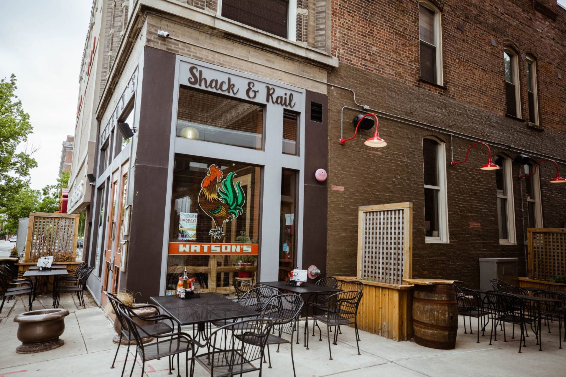 The corner of a brick building with black patio tables and chairs in front of it. On one of the windows is a red and green painted rooster, and on the building above it says Shack & Rail in black script.