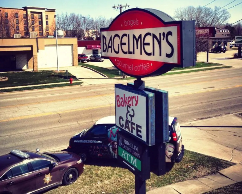 A picture taken from above of two cars and a sign in front of a two-lane street. the sign is a red oval with a white strip through the middle. it says Bagelmen's