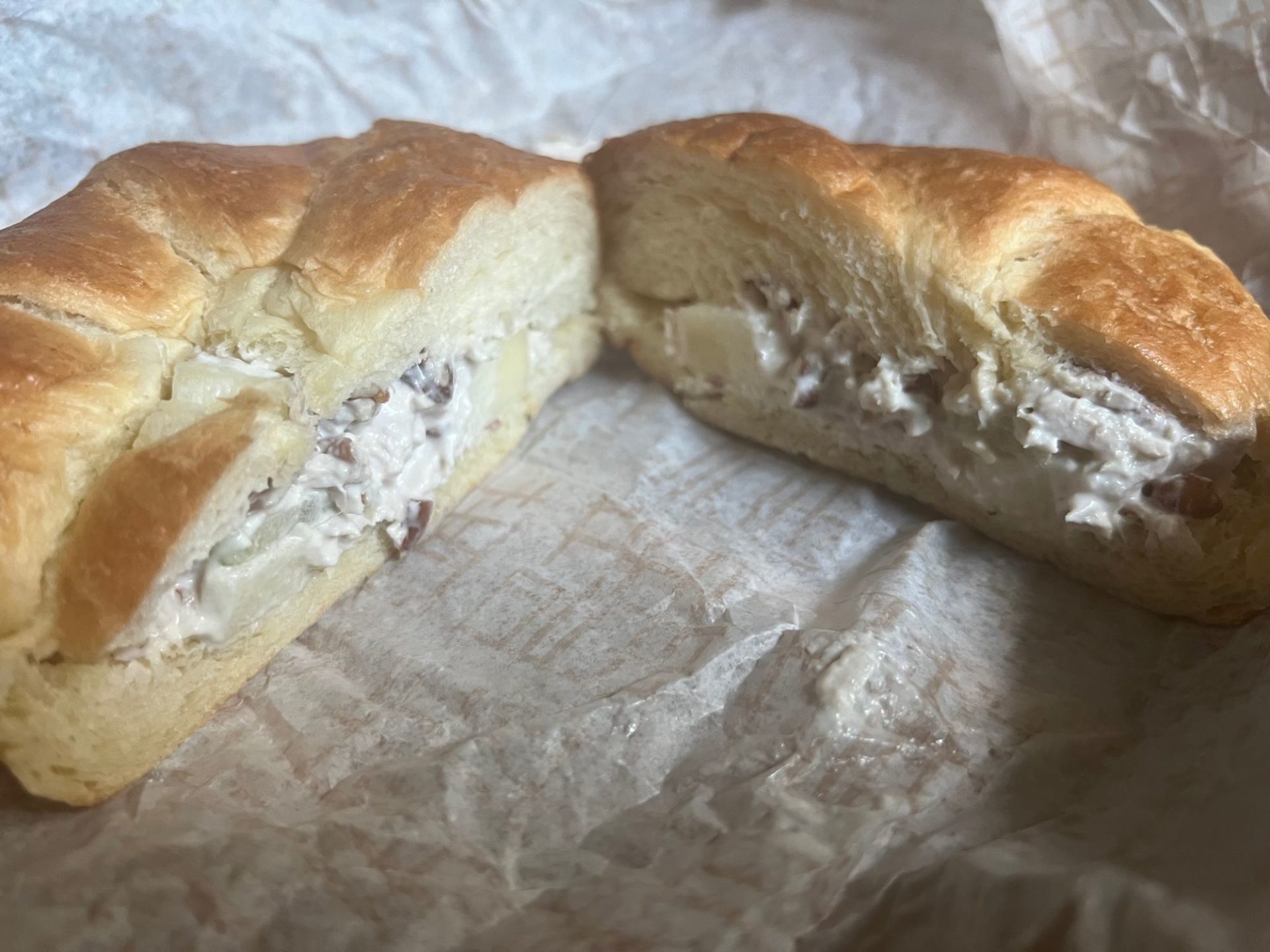 A flaky croissant sandwich with turkey salad filling sits on a crinkled white wrapper. It's cut in half and displayed at an angle.