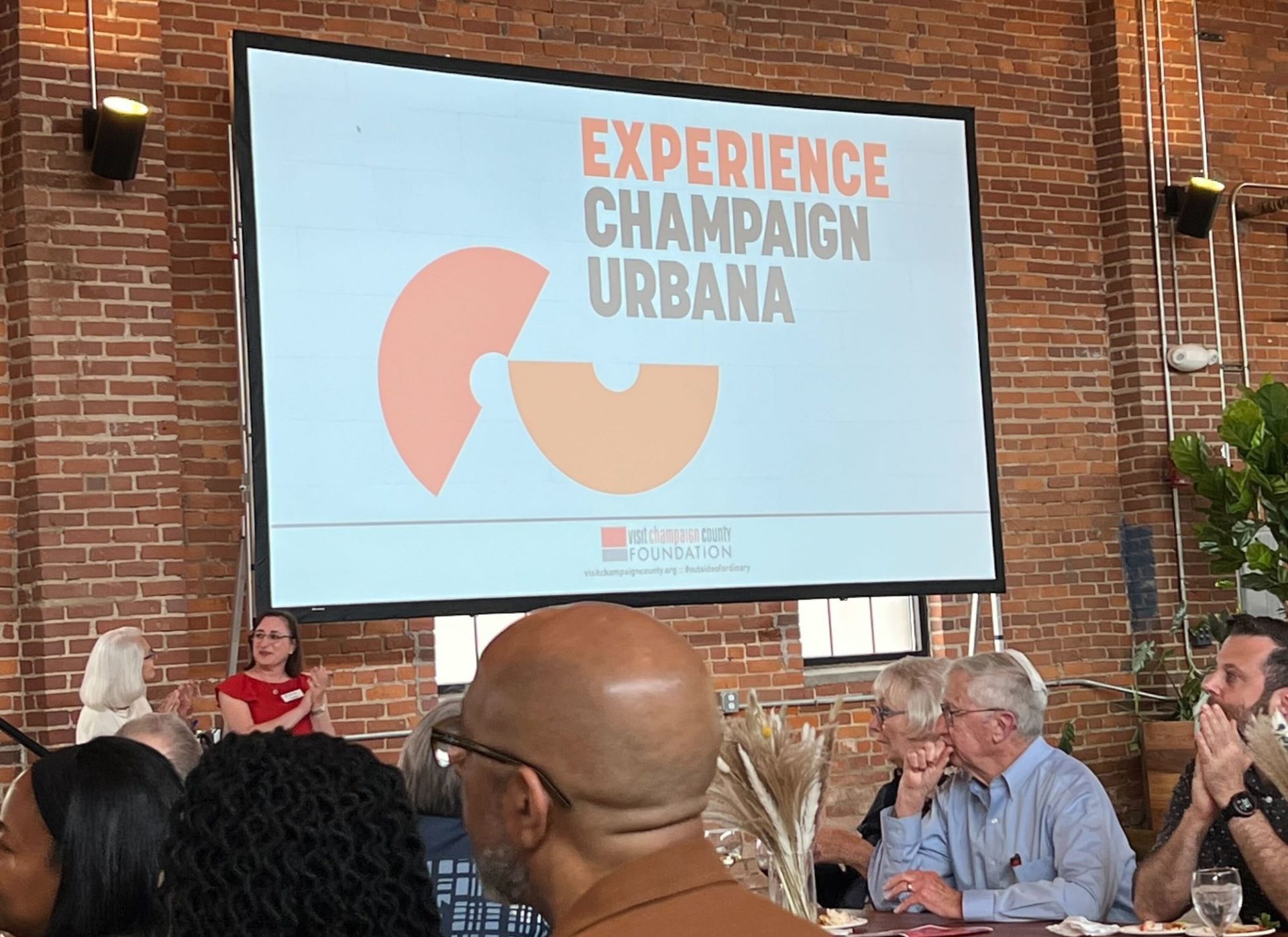 A large screen attached to a brick wall. It has a white background, and is displaying two orange semi circles and Experience Champaign Urbana in block lettering.